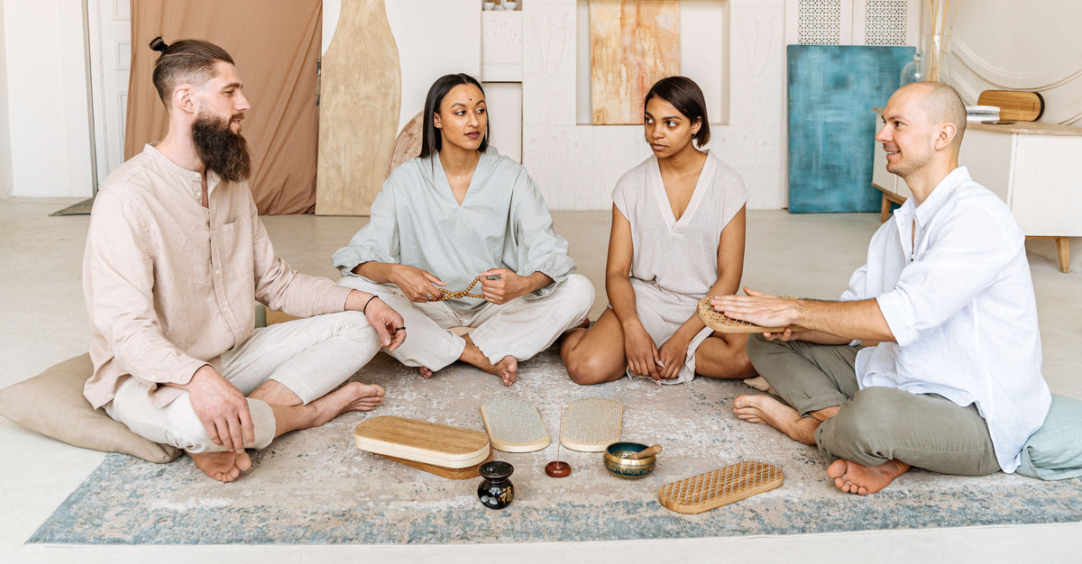 Four people sitting cross-legged on a blue patterned rug in a bright, modern room, gathered around small tabletop instruments including wooden blocks, a bowl, and a drum.